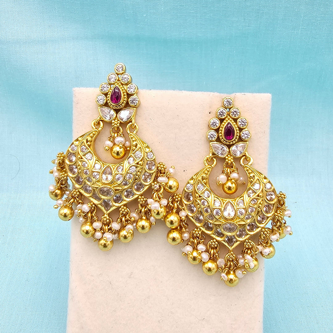 A pair of ornate gold-toned Indian jhumka earrings with clear stones, small white bead drops, and a central red stone, displayed on a cream stand against a light blue fabric background.